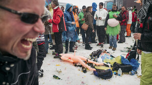 Spaß beim Apres Ski - Ein Mann lacht, es wird getrunken und die Leute stehen rum und ein Sexpuppe liegt im Schnee