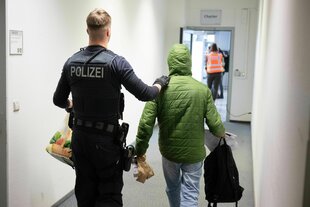 Ein Polizeibeamter in Uniform begleitet einen Mann in einem grünen Anorak, er hat die Hand suaf seine Schulter gelegt - sie gehen einen Flur ntlang