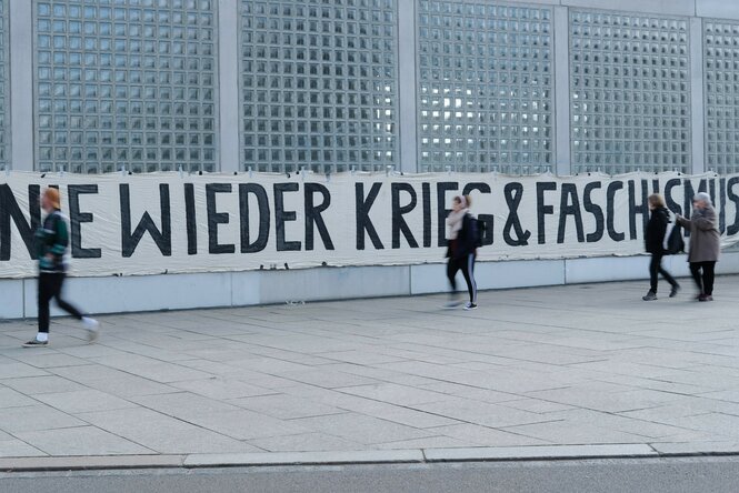 Ein Banner mit der Aufschrift "Nie wieder Krieg & Faschismus" ist an der Fassade einer S-Bahnstation angebracht.