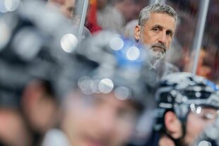 Unscharf im Vordergrund behelmte Eishockeyspieler, im Hintergrund scharf zu sehen das Gesicht von Trainer Mark French