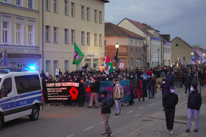 Ein Demonstrationszug, angeführt von einem Polizeifahrzeug, zieht durch Teltow am frühen Abend. Ganz vorne ein Banner mit der Aufschrift "Kampf dem Faschismus"