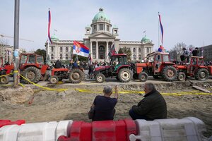 Eine Reihe von Traktoren verdeckt eine demonstrierende Menge vor dem Parlament in Belgrad. Im Vordergrund eine Art Sandgrube, in der zwei Männer sitzen