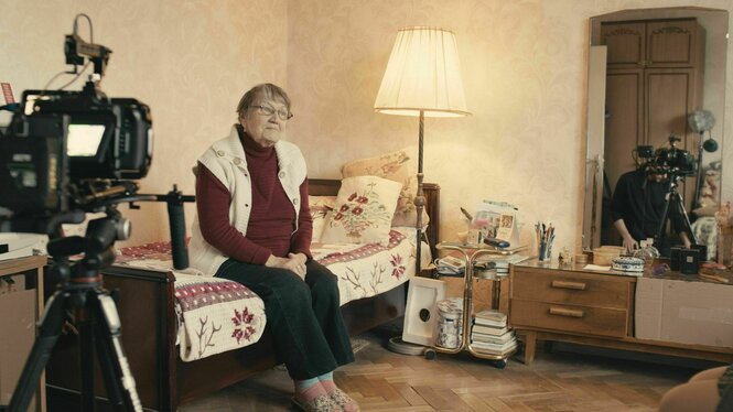 Eine Frau sitzt auf einem Bett, daneben stehen eine Lampe und eine Filmkamera