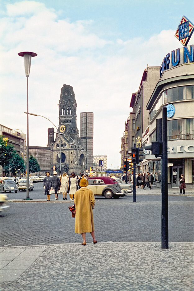 Rückenansicht einer Frau im eleganten gelben Mantel, die an der Straße steht, vor ihr die Gedächtniskirche, der Kurfürstendamm ist gepflastert und noch nicht asphaltiert