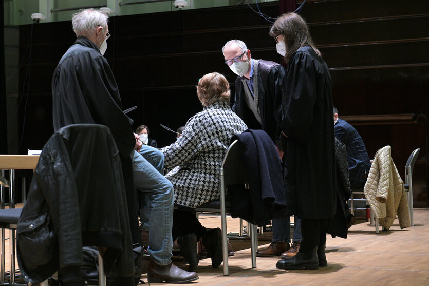 Ulrike B. sitzt auf einem Stuhl und unterhält sich während des Strafprozesses im Bremer Konzerthaus Glocke 2021 mit drei Anwält*innen; die drei Verteidiger*innen tragen Anwaltsroben und FFP-2-Masken, B. selbst ist nur von hinten zu sehen