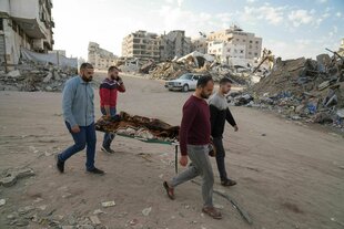 Vier Männer tragen eine Bahre, auf der ein in einen Teppich gewickelter Leichnam liegt, durch eine Ruinenlandschaft