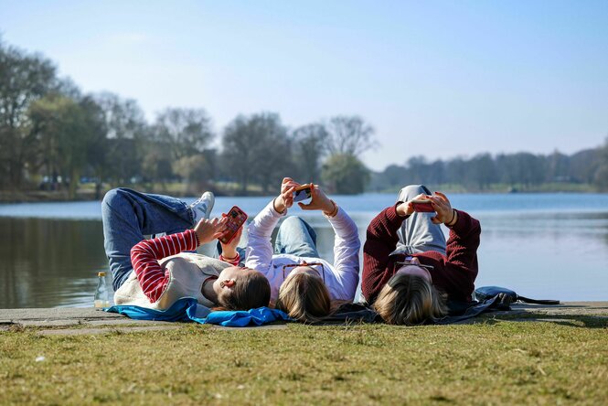 Drei junge Frauen liegen auf einer Wiese und schauen auf ihre Handys.