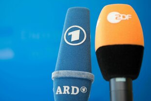 Die Mikrofone von ARD und ZDF sind vor einer blauen Wand nebeneinander aufgestellt