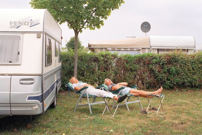 Ein Mann und eine Frau liegen vor einem Campingwagen auf zwei Liegestühlen und schlafen am helllichten Tag