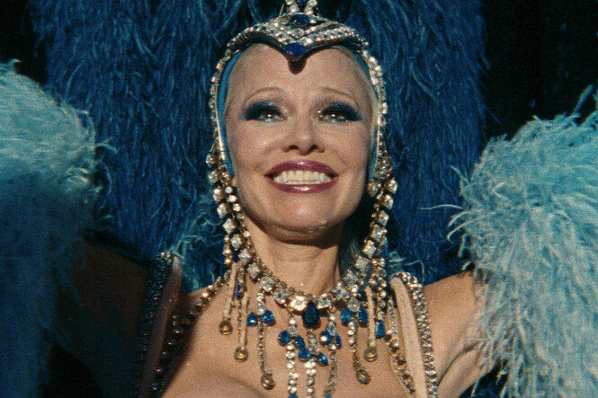 Pamela Anderson in der Hauptrolle des Films "The Last Showgirl" strahlt in Kostum mit blauen Engelsflügeln gezwungen in die Kamera