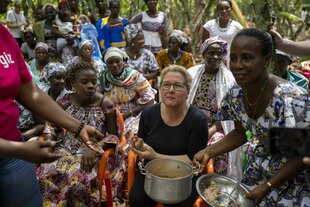 Svenja Schulze, umringt von Frauen - sie probiert eine lokal gekochte Fischsuppe auf einer Kakaoplantage