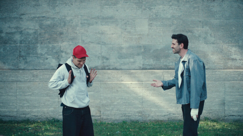 Ein Filmstill des Films "Sad Jokes" vom Regisseur Fabian Stumm. Zwei Männer stehen vor einer grauen Häuserfront. Der rechte wendet sich dem linken zu, der mit beiden Händen lässig abwinkt.