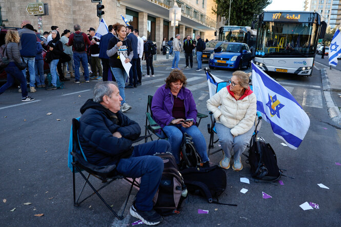 Menschen besetzen eine Straße bei einer Kundgebung gegen den israelischen Premierminister Benjamin Netanjahu und seine Regierung, sie sitzen in drei Stühlen und einer israelischen Flagge auf der Straße