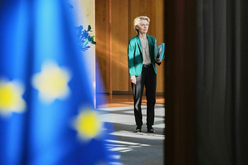 Ursula von der Leyen kommt zur wöchentlichen Sitzung , sie steht vor einer holzgetäfelten Wand, im Vordergrund ist unscharf der Teil einer EU-Flagge zu erkennen