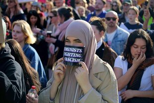 Eine junge Frau mit Kopftuch hält sich auf einer Protestaktion am Frauentag ein Schild vor das Gesicht: Feminists Fighting Fascism