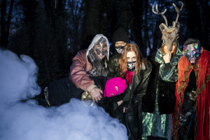 Das Foto zeigt im Wald stehen Personen. Es ist Nebel im Vordergrund (aus einer Nebelmaschine). Die Personen haben Masken auf. Die Masken sind unterschiedlich von pinker Wollmaske, Geweih oder moderne Prismamaske.