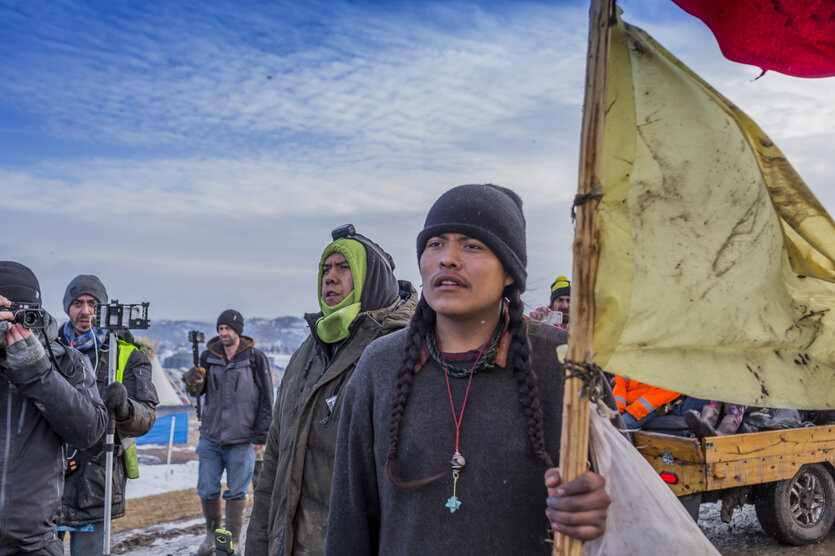 Protestierende in einer kalten Winterlandschaft. Ein Mann mit langen Zöpfen und Mütze hält eine Fahne in der Hand