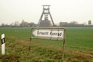 Ein Hinweisschild, Aufschrift „Schacht Konrad“, im Hintergrund ein Förderturm