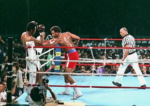 George Foreman verpasst Muhammad Ali einen Körpertreffer im Ring, Ali hängt in den Seilen. Der Schiedsrichter läuft in einiger Entfernung im Ring.