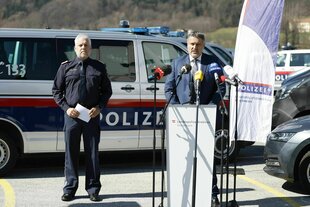 Zwei mittelalte Männer, einer in Uniform, der andere im Anzug, stehen bei Sonnenschein vor Polizeiautos, der Mann im SAnzug spricht in ein Mikrofon, der andere schaut ernst und hält Notizen in der Hand