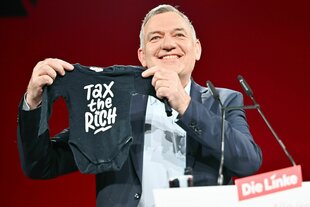 Van Aken hält einen Strampler mit der Aufschrift "Tax the Rich"