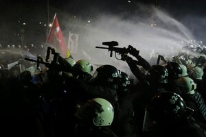 Gepanzerte Polizeikräfte gehen auf einer nächtlichen Straße gegen Demonstranten vor, im Hintergrund die Strahlen von Wasserwerfern