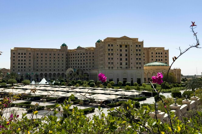 Blick auf ein großes Luxushotel mit gepflegtem parkartigen Garten und Springbrunnen, im Vordergrund überdachte Parkplätze