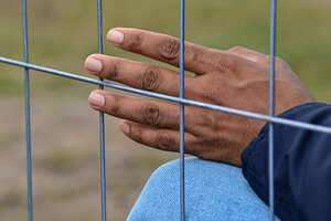 Nahaufnahme: Die Hand einer geflüchteten Person scheint beinahe behutsam einen Bauzaun anzufassen