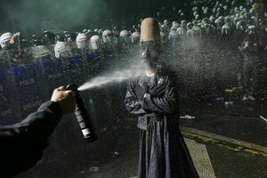 Eine große Kanne Pfefferspray wird einer demonstrierenden Person mit Gasmaske und traditionellem Filzhut direkt und aus nächster Nähe in gesicht gepfeffert. Die Person steht vehement, widerspenstig trotzend mit den Armen verschränkt still.