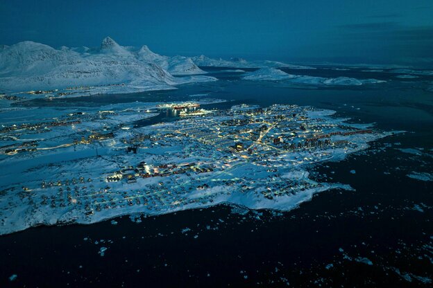 Luftbild von Nuuk, der Hauptstadt Grönlands, in der Dämmerung. Die Stadt zeichnet sich durch die beleuchteten Straßen und vorallem den Hafen ab, im Kontrast zu dem sie umgebenden Schnee und Eis.