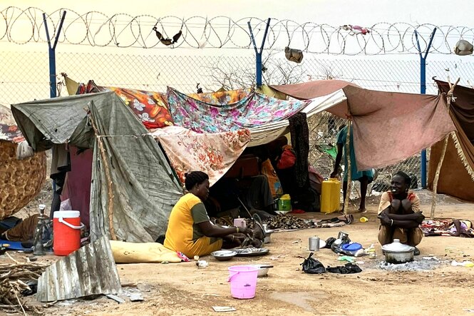 Zwei Frauen sitzen vor Zelten auf dem Boden und bereiten Essen zu, hinter ihnen stehen provisorische Zelte mit Tüchern, die an einem Stacheldrahtzaun befestigt sind