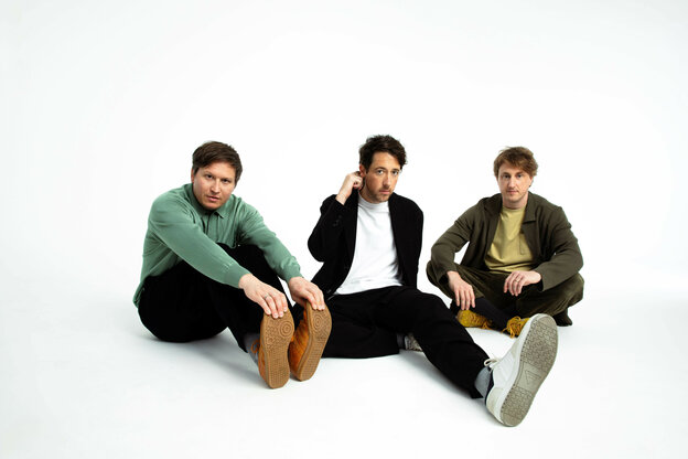 Ein Foto der drei Bandmitglieder der Band „The Wombats“. Die „Jungs“ sitzen auf dem Boden in einer weißen Studiohohlkehle.