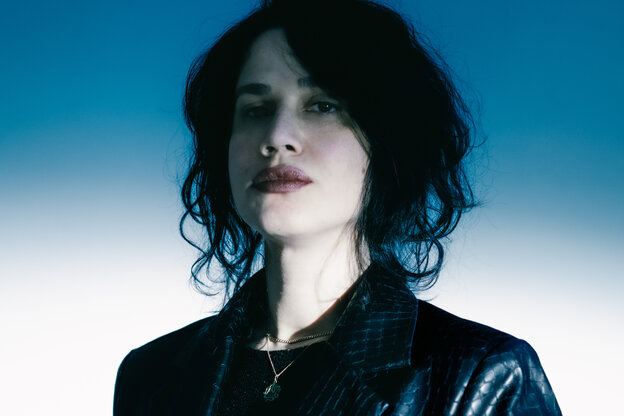Portraitfoto von Hüma Utku. Die Musikerin steht vor einem blau-weißem Hintergrund und trägt eine schwarze Lederjacke und eine Kette. Sie hat schwarze, kinnlange Haare und trägt roten Lippenstift.