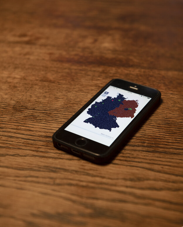 Fotoarbeit von Göran Gnaudschun. Auf einer braunen Holztischplatte liegt ein Smartphone, dessen Bildschirm eine Deutschlandkarte mit nach Farben markierten Wahlergebnissen anzeigt.