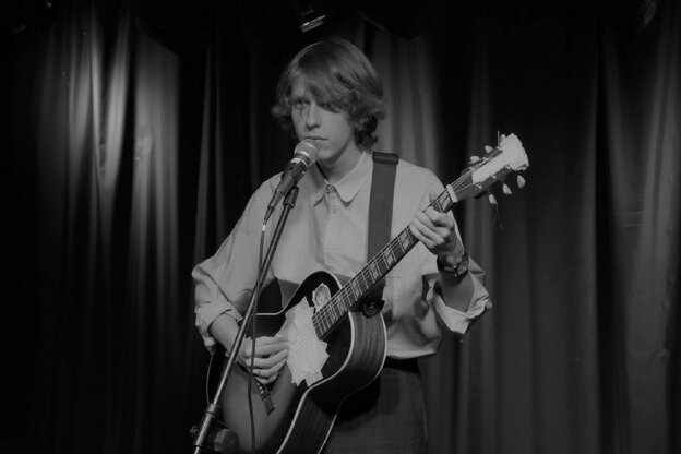 Szene aus „The Swan Song of Fedor Ozerov“: Das Filmstill zeigte eine Szene in schwarz-weiß, ein junger Mann steht mit einer Gitarre vor einem Mikrofon. Er trägt ein Hemd. Die Öffnung der Gitarre ist mit Klebeband zugeklebt.
