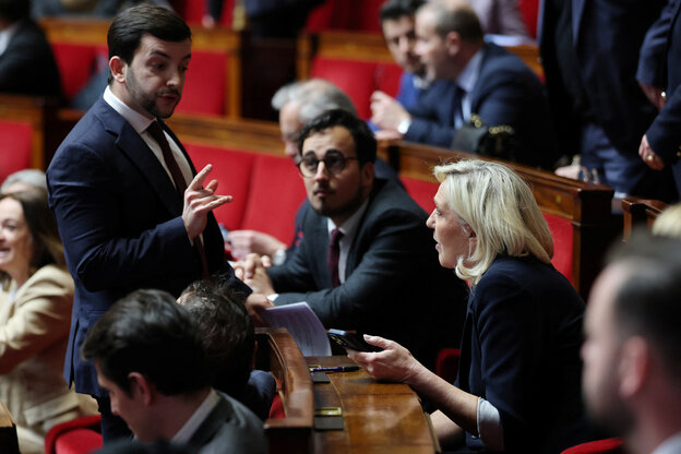 Marine Le Pen sitzt in der Nationalversammlung, hält ein Handy, ein Mann vor ihr gestikuliert heftig