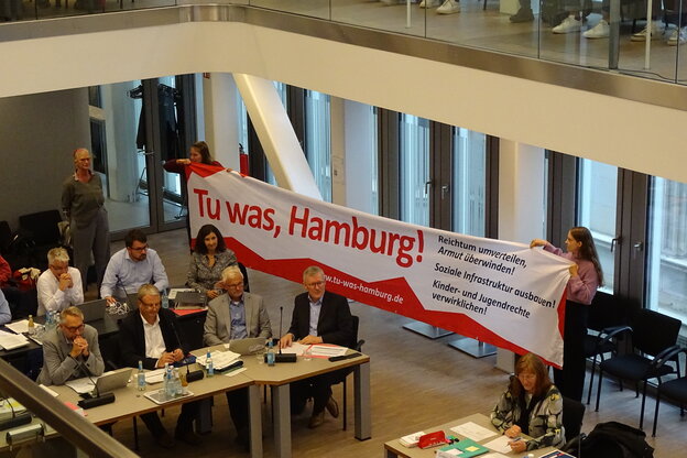 Ein Raum mit Menschen, einige sitzen, zwei halten ein Transparent mit der Aufschrift 