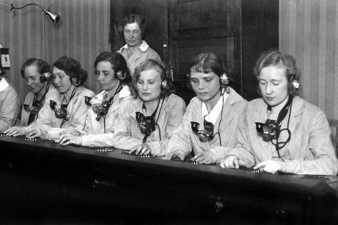 Telefonistinnen sitzen mit Kopfhörer an einem Tisch mit Schalthebeln - schwarz-weiss Aufnahme aus den 30er Jahren