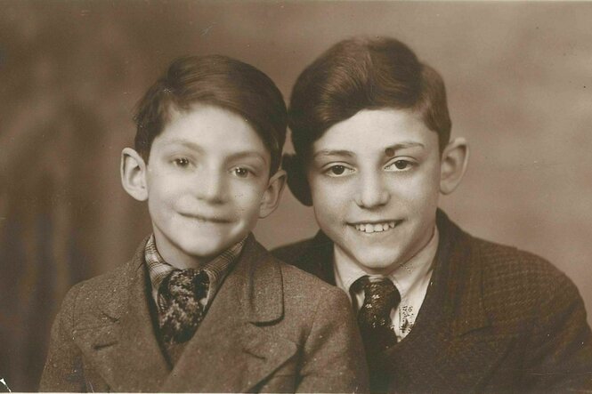 Schwarz-Weiß-Foto von zwei Jungen von der Brust aufwärts. Sie sitzen eng beisammen, lächeln für das Foto. Beide haben dunkle Haare und einen Seitenscheitel, tragen Anzug und Hemd.