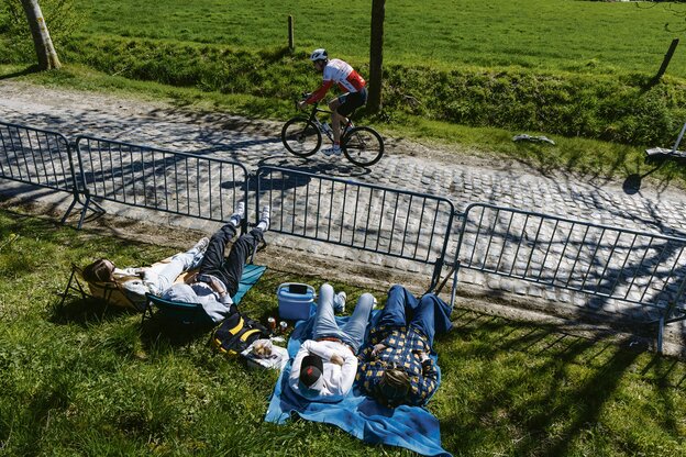 Ein Radsportler fährt auf Kopfsteinpflaster, während Fans auf Campingstühlen und mit Picknick-Equipment auf der Wiese sitzen