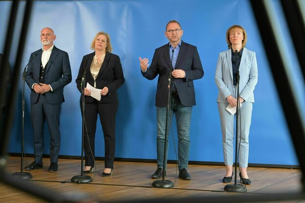Vier Personen stehen nebeneinander vor Mikrofonen und schauen ernst