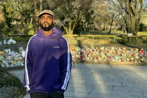 Ein Mann in violetter Trainingsjacke steht vor einer Gedenkstätte in einem Park