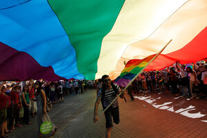 Man sieht: eine Pride Parade mit vielen Menschen. Über ihnen schwebt eine großen Regenbogenfahne.