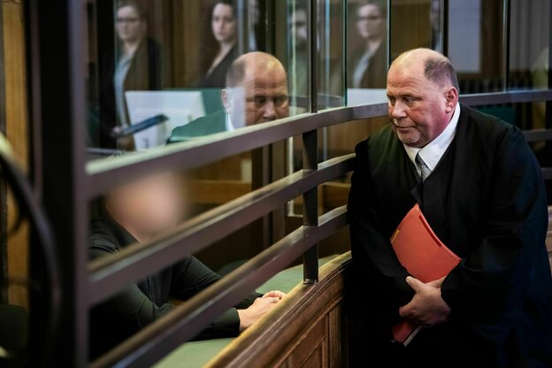Ein Anwalt im Gericht. Hinter einer Galsschreibe sitzt der Angeklagte