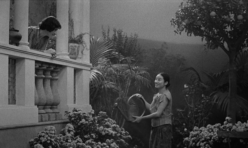 Das schwarz-weiße Filmstill zeigt Cláudio da Silva als Timothy Sanders auf einem Balkon und Lang Khê Tran als Ngoc im Garten davor