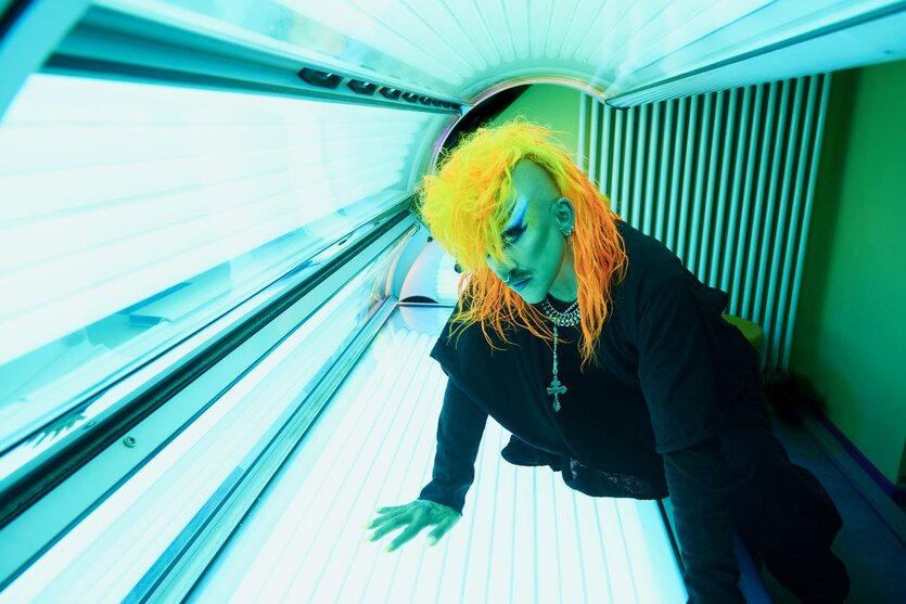 Eine Person in dunkler Kleidung, mit orangen Haaren, Schnurrbart und ausladender Schminke