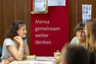 Einige Frauen unterhalten sich bei dem Jahrestreffen des Hochbegabten-Vereins Mensa im Jahr 2024; hinter dem Tisch hängt ein Banner mit dem Aufspruch "Mensa gemeinsam weiter denken"