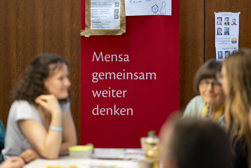 Einige Frauen unterhalten sich bei dem Jahrestreffen des Hochbegabten-Vereins Mensa im Jahr 2024; hinter dem Tisch hängt ein Banner mit dem Aufspruch "Mensa gemeinsam weiter denken"