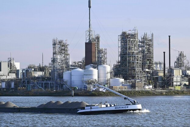 Eine Industrielandschaft an einem Fluss, es schiebt sich eine Lastkahn vopn links mit dem Bug ins Bild