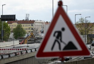 Ein Verkehrsschild weist auf Bauarbeiten hin, im Hintergrund sind Bauarbeiten an einer Brücke zu sehen